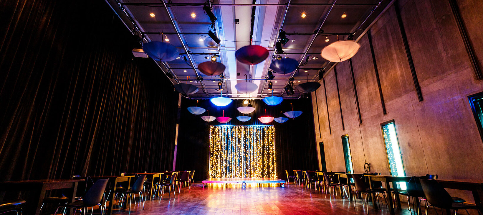 A large room is dimly lit with chairs and tables on either side of the floor, multi coloured umbrellas hanging from the ceiling and a glittery platform stage is at the end of the room.
