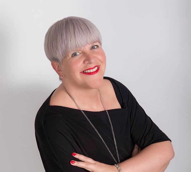 Image shows Sally-Anne smiling at the camera with her arms crossed. They are wearing a black dress, a long necklace and red lipstick. They have short white/grey hair and are standing in front of a white background.